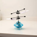 A small hovering toy helicopter with a transparent light blue body, black rotor arms, and white blades, photographed on a plain light surface against a light wall.
