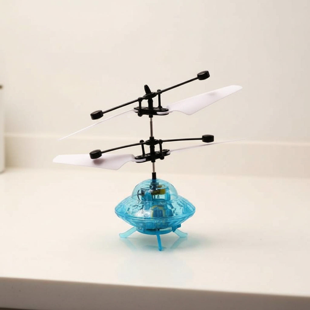 A small hovering toy helicopter with a transparent light blue body, black rotor arms, and white blades, photographed on a plain light surface against a light wall.