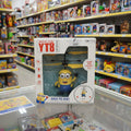 A boxed toy helicopter featuring a Minions character, displayed on a glass counter inside a toy store. The packaging is white with red and blue text and shows the product through a clear window.