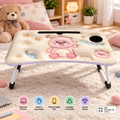 A portable folding lap desk with a cute cartoon bear design, shown on a carpeted floor in a cozy room setting. The desk features a cream-colored surface with a pink bear graphic, a built-in cup holder, a phone/tablet slot, and a circular cutout.