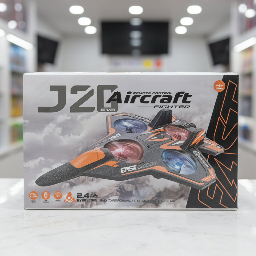 A boxed remote control aircraft toy labeled 'J20 Remote Control Aircraft Fighter' sits on a light countertop in a blurred indoor retail or showroom environment. The box shows a black and orange jet design with cockpit windows and product features along the bottom.