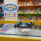 A blue and black quadcopter drone displayed on a clear acrylic stand on a toy store counter, with colorful toy shelves and an 'Adventure Toys' sign in the blurred background.