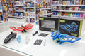 A WRC Sky drone kit displayed on a glass tabletop in a toy store, showing the red and blue drones, a black remote controller, propellers, cables, and two product boxes.