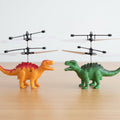 Two toy helicopters mounted on dinosaur figures, one orange with a pink back and one green with dark green spikes, displayed on a light wooden surface against a white background.
