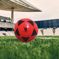 Red soccer ball with black pentagonal patterns on a grass field, likely in a stadium.